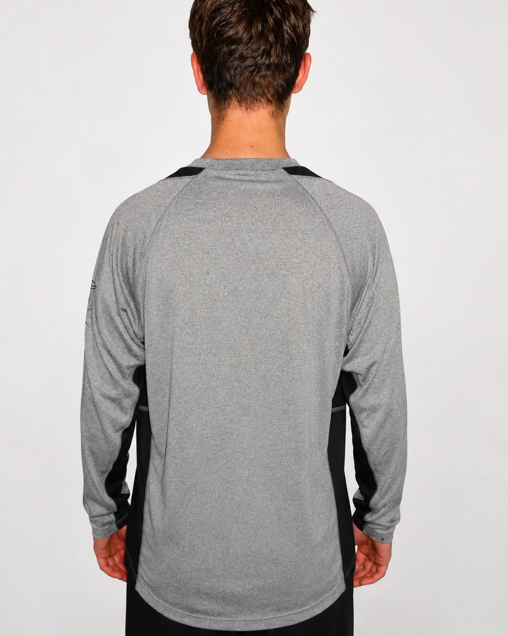 A man viewed from the back wearing a heather gray long-sleeve athletic shirt with black side panels and black shoulder detailing, against a plain light gray studio background.