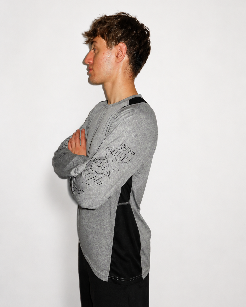 A man in profile wearing a heather gray long-sleeve athletic shirt with black side panels and a black mountain sleeve graphic, posed against a plain light gray background.