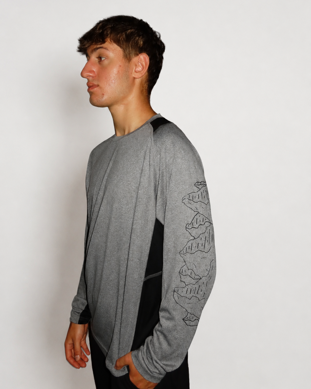 A man wearing a heather gray long-sleeve shirt with black side panels and a black mountain line-art graphic on the sleeve, shown in profile against a plain light gray background.